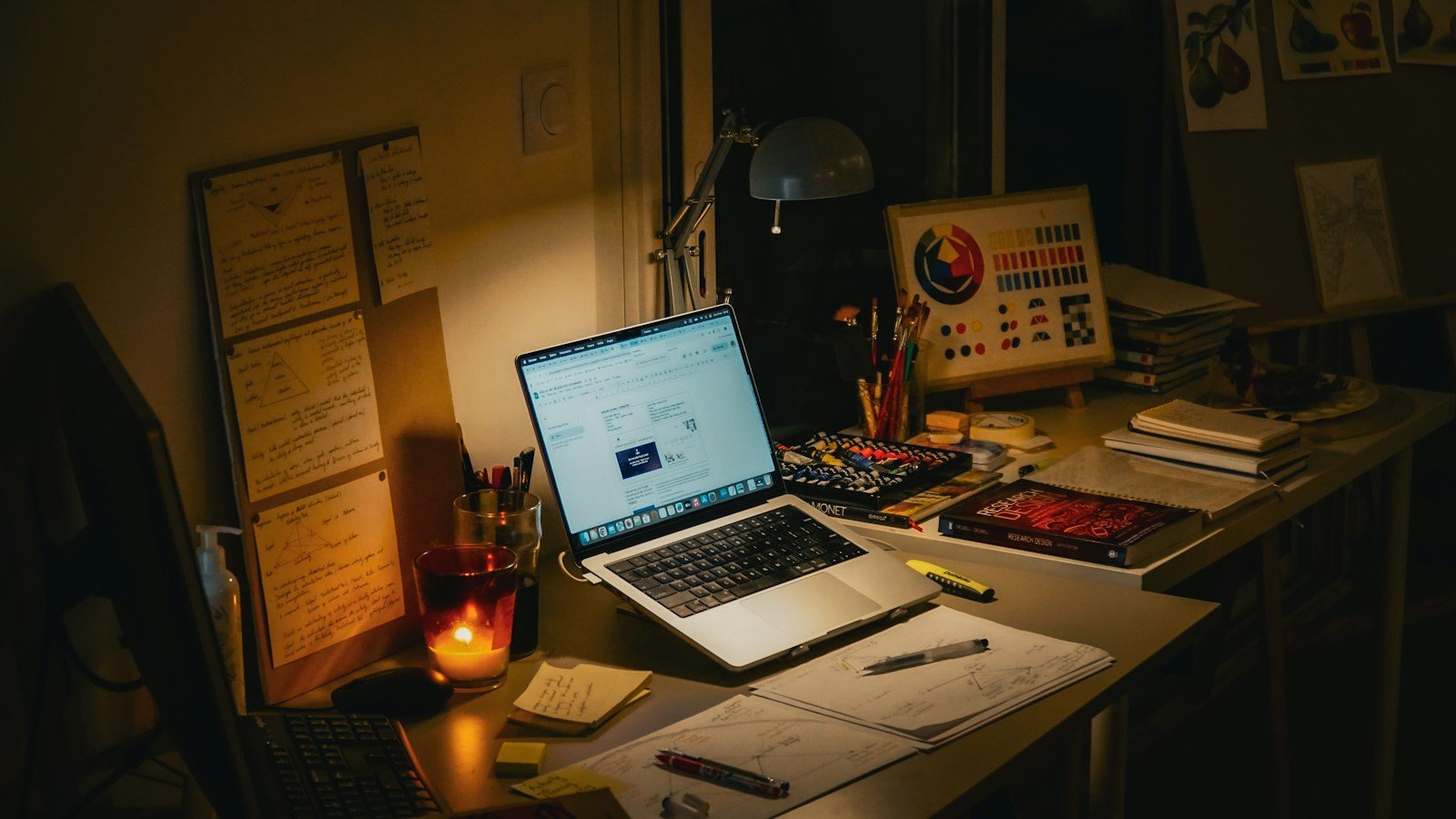 夜の机に、ノートパソコンと手書きのメモ、本、灯したキャンドルが置かれている